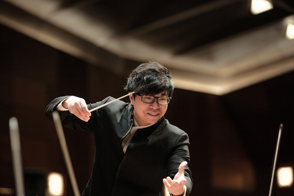Kahchun Wong, a man wearing smart black attire, conducting at the Queen Elizabeth Hall, smiling in a performance.